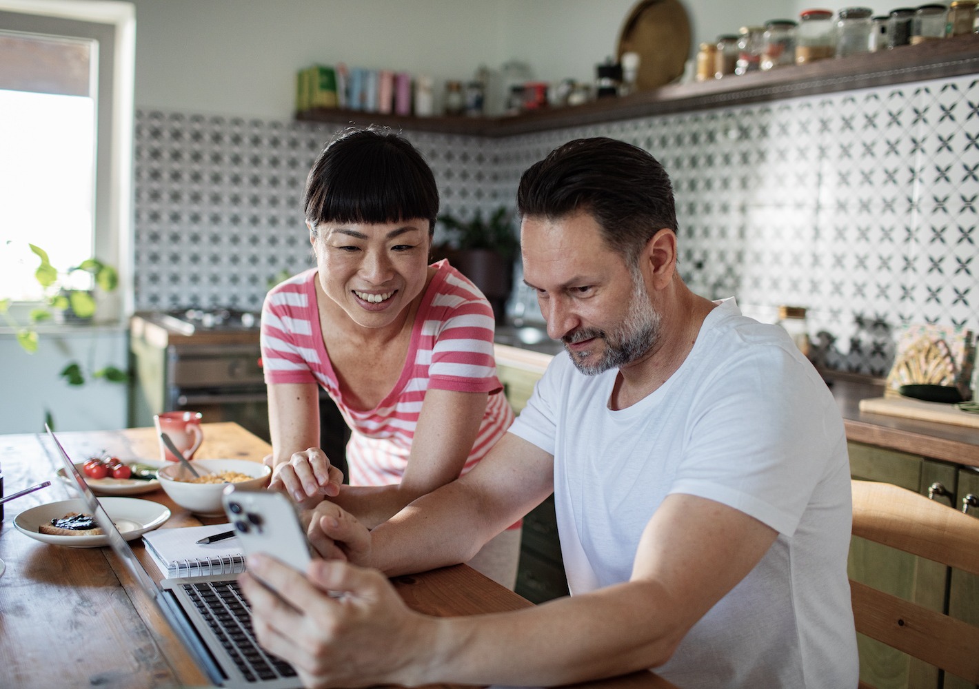 Couple using phone and laptop at kitchen table during breakfast